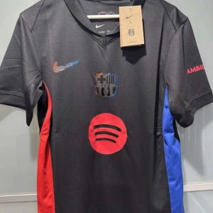 FC Barcelona svart matchtröja Nike - Säljer en svart FC Barcelona fotbollströja från Nike med klubbmärke och sponsorlogga på bröstet. Tröjan har röda och blå detaljer på sidorna, korta ärmar och tryckt text på ärmen. Perfekt för dig som älskar Barca! Finns alla storlekar 