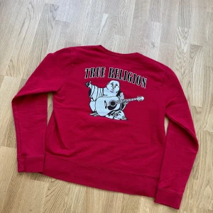 Röd sweatshirt från True Religion size M - Röd sweatshirt från True Religion med logga framtill och stort tryck med buddha och gitarr på ryggen. Rund halsringning och långa ärmar. Skön och avslappnad passform, perfekt till vardags.