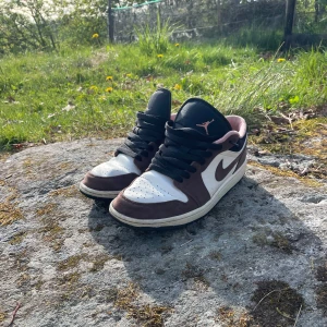 Nike Air Jordan 1 Low mocha  - Säljer ett par Nike Air Jordan 1 low mocha med brun mocka, vita partier och svarta detaljer. Skorna har svart snörning och svart sula. Perfekta för dig som gillar streetwear och unika sneakers.OBS Det finns en defekt som man ser på bild 5.