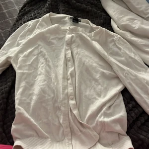 Vit kofta från H&M - Säljer en klassisk vit kofta från H&M. Koftan har långa ärmar, knappar framtill och ribbade muddar vid ärmslut och nederkant. Perfekt att slänga över en topp eller klänning för en enkel och stilren look.Storlek L men sitter som Xs