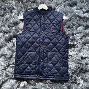Blå quiltad väst från Polo Ralph Lauren - Snygg blå quiltad väst från Polo Ralph Lauren med knappar framtill. En liten logga på bröstet. Perfekt för lager-på-lager-stil. Västen är i storlek 14-16 år. Mycket bra skick inga defekter