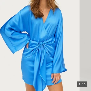 Blå omlottklänning i satin - Säljer en snygg blå omlottklänning i satin med lång ärm och stor knytning i midjan. Klänningen har v-ringning och en kort längd som passar perfekt till speciella tillfällen ifrån gina