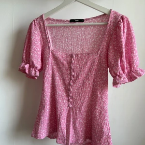 Rosa blommig blus med puffärmar från Kook - Supersöt rosa blus med små vita blommor, puffiga kortärmar med volang och knappar framtill. Fyrkantig ringning och lätt rynkat tyg ger en romantisk vibe. Perfekt för dig som gillar feminina detaljer!