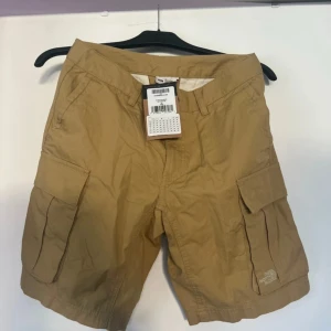 Khaki cargoshorts från The North Face - Snygga khaki cargoshorts från The North Face med flera praktiska fickor på sidorna och bak. Klassisk cargomodell med normal passform och diskret logga på benet. Perfekta för sommaren och tillverkade i slitstark bomull. Köptes för 600 kr och är helt oanvänd. 