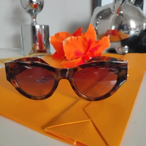 Bruna solglasögon från Saint Laurent - Snygga solglasögon från Saint Laurent med brunmelerat sköldpaddsmönster och breda bågar. Modellen har en klassisk cat-eye form och mörktonade linser. Perfekt accessoar för att lyfta din stil.