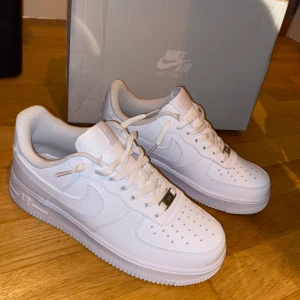 Nike Air Force 1 - Klassiska helvita Nike Air Force 1 '07 sneakers med låg profil och ikonisk design. Skorna har vit ovandel i skinn, perforerad tå och platt sula. Perfekta för dig som gillar stilrena och tidlösa sneakers.