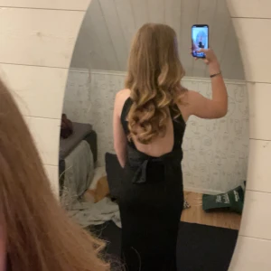 Svart långklänning med öppen rygg och rosett - Säljer en elegant svart långklänning med öppen rygg och stor rosett bak. Klänningen har en tight passform och är ärmlös, perfekt för bal eller fest🎊