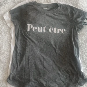 Grå t-shirt med Peut-être-tryck - Snygg grå t-shirt med vit text 'Peut-être' framtill. Klassisk rund halsringning och korta ärmar. Perfekt för en chill och avslappnad stil. Mjuk och bekväm, passar till jeans eller shorts.