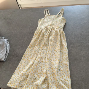 Blommig sommarklänning från Zara - Supersöt långklänning från Zara med smala axelband som korsas i ryggen. Klänningen är vit med små gula blommor och har en smockad överdel. Perfekt för varma dagar och ger en fräsch och somrig vibe.