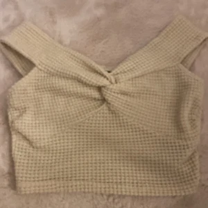 Beige croppad stickad topp - Superfin croppad topp i beige stickat material med twistad detalj framtill. Toppen har breda axelband och en snygg, fyrkantig form. Perfekt att styla med höga jeans eller kjol för en chill och trendig look.