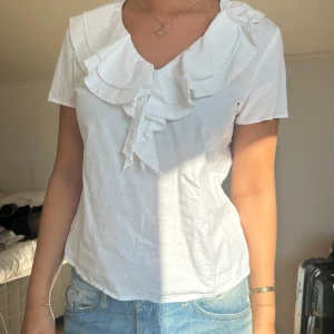 Vit blus med volangkrage - Supersöt vit blus med korta ärmar och stor volangkrage som ger en romantisk vibe. Blusen har v-ringning och är gjord i mjuk bomull, perfekt till jeans eller kjol. Volangdetaljerna gör den extra speciell och den är lätt att styla för en trendig look.