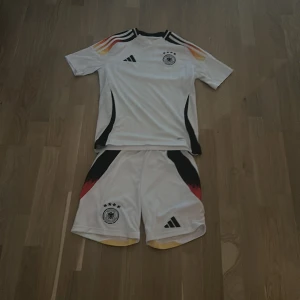 Tysklands matchställ Adidas vit - Säljer ett Tysklands fotbollsställ från Adidas. Tröjan är vit med svarta detaljer och korta ärmar, har Tysklands landslagsmärke och Adidas-logga på bröstet. Axelpartiet har ränder i svart, rött och gult. Shortsen är vita med svarta och röda detaljer samt landslagsmärke och Adidas-logga. Materialet är lätt och andas.
