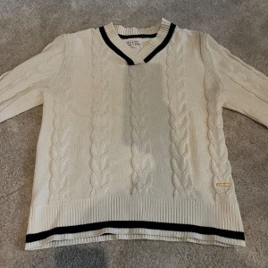 Vit kabelstickad tröja från Marine - Vit kabelstickad tröja från Marine som aldrig är använd med svarta ränder vid v-ringad krage och nederkant. Tröjan har lång ärm, ribbade muddar och en snygg badge i guld på ena sidan. Perfekt för en preppy look och passar till många outfits.