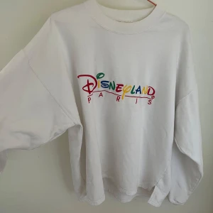 Vit Disneyland Paris sweatshirt S/M - Vit sweatshirt från Disneyland Paris med broderad färgglad logga på bröstet. Oversized passform, rund hals och långa ärmar. Tillverkad i mjuk bomull och polyester. Lite missfärgad vid kragen. 