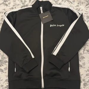 Svart track jacket från Palm Angels - Svart track jacket från Palm Angels med vita ränder längs ärmarna och vit dragkedja. Jackan har ståkrage, ribbade muddar och logotypen tryckt på bröstet. Perfekt för en sportig och trendig look.