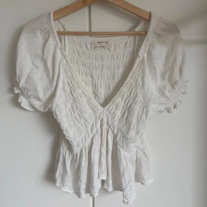 Vit blus från Urban Outfitters XS - Supersöt vit blus från Urban Outfitters med puffiga korta ärmar och v-ringning. Blusen har smockad struktur och lätt volang nertill💕 storlek xs men är stretchig så passar mig som har S/M också 🥰