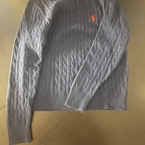 Mörkblå kabelstickad tröja  - Kabelstickad mörkblå tröja från med klassisk orange logga broderad på bröstet. Tröjan har rund halsringning, långa ärmar och ribbade muddar. Perfekt för dig som gillar en stilren och tidlös look. OBS INTE ÄKTA
