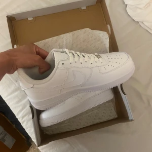 Nike Air Force 1 helvita sneakers - Klassiska Nike Air Force 1 i helvitt skinn med låg profil och perforerad tå. Snygg och clean look med vit sula, snörning och ikoniska swoosh-loggan på sidorna. Perfekta för dig som vill ha en tidlös sneaker med streetkänsla.