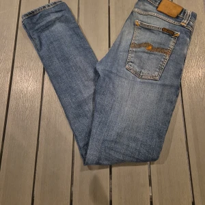 Nudie Jeans - Säljer ett par klassiska blå skinny jeans från Nudie Jeans med snygga kontrastsömmar och ikonisk brodyr på bakfickorna. Jeansen har normal passform, låg midja och är tillverkade i mjukt bomullsjeans. Perfekta för dig som gillar en smal siluett och stilren look. Jag har lagat jeansen på ett ställe 