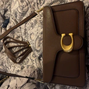 Brun axelväska från Coach - Säljer en stilren brun axelväska från Coach med grovkornigt skinn och en stor guldfärgad C-detalj på framsidan. Väskan har ett kort axelband och även en lång (den är inte äkta) VÄSKAN ÄR INTE ÄKTA 