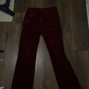 Vinröda velourbyxor från Juicy Couture - Mysiga vinröda mjukisbyxor från Juicy Couture i mjuk velour med snörning i midjan och fickor fram. Bak på byxan står det 'Juicy' i glittriga stenar. Perfekta för en chill och trendig look.