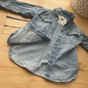 Ljusblå jeansskjorta från H&M - Snygg ljusblå jeansskjorta från H&M med klassisk krage, tryckknappar och två bröstfickor med lock. Skjortan är tillverkad i bomull och har långa ärmar. Perfekt för en cool och avslappnad look.
