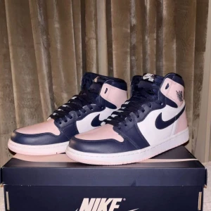 Nike Air Jordan 1 High rosa/marinblå - Nike Air Jordan 1 High i en clean färgkombo av ljusrosa, marinblått och vitt. Skorna har klassisk snörning, rund tå och är tillverkade i skinn. Snygga detaljer med kontrasterande paneler och ikonisk Nike-logga på sidan. Perfekta för dig som vill sticka ut med din sneakerstil.