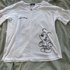 Vit Mickey Mouse t-shirt från Sheilay - Vit t-shirt från Sheilay med svart tryck av Mickey Mouse på framsidan och texten 'Mickey Mouse' vid bröstet. T-shirten har korta ärmar, rund halsringning och är gjord i ett tunt, mjukt bomullstyg. Perfekt för dig som gillar Disney och vill ha en chill look.