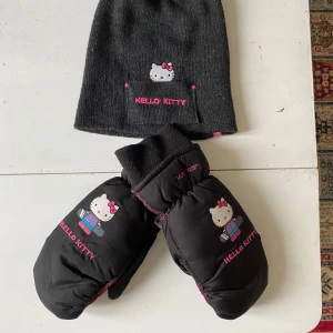 Svarta Hello Kitty vintervantar och mössa - Svarta tumvantar från Hot Paws med Hello Kitty-tryck och broderad text i rosa. Insidan har rosa mönster och muddar i svart fleece. Perfekta för kalla vinterdagar och för dig som älskar Hello Kitty. Jag säljer även mössan till. Allt för 80 kr.
