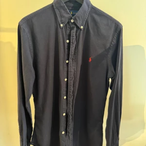 Mörkblå skjorta från Ralph Lauren - Klassisk mörkblå skjorta från Ralph Lauren med lång ärm och knappar hela vägen. Skjortan har en röd broderad logga på bröstet och button-down krage. Tillverkad i mjuk bomull och passar perfekt till jeans eller chinos.