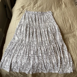 Vit långkjol med svart prickigt från Vila - Superfin långkjol från Vila i vitt med svarta prickar. Kjolen har en luftig och flowy passform med flera volanger och resår i midjan. Perfekt för dig som gillar en avslappnad men ändå trendig stil.