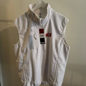 Vit väst från Clique med dragkedja Smirnoff Ice - Vit väst från Clique branded Smirnoff Ice med hög krage och hel dragkedja framtill. Västen har två sidofickor med dragkedja och en bröstficka. Materialet är slätt och känns sportigt, perfekt för lager-på-lager. Röd logga på bröstet ger en snygg kontrast.