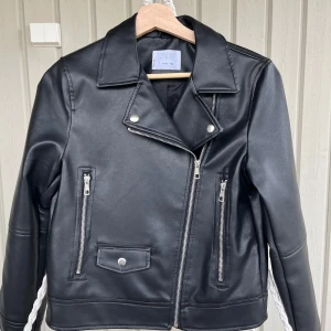 Svart skinnjacka från Zara, stl 152 - Snygg svart skinnjacka från Zara med klassisk bikerstil. Jackan har silvriga dragkedjor, bred krage och två fickor framtill. Materialet är syntetiskt skinn och jackan har raka, långa ärmar. Perfekt för dig som vill ha en cool och tidlös look.