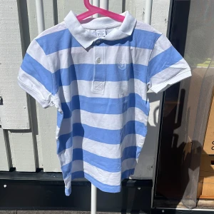 Randig pikétröja från Lindex blå/vit - Snygg pikétröja från Lindex i storlek 146/152. Tröjan har breda blå och vita ränder, klassisk krage och knäppning med två knappar framtill. Tillverkad i mjuk bomull och har korta ärmar. Perfekt för en fräsch och sportig look i sommar.