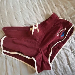 Vinröda shorts från Hollister - Snygga vinröda shorts från Hollister med vit snörning i midjan och vita kanter. Klassisk sportig look med broderad Hollister-logga och blå mås på ena benet. Perfekta för sommardagar eller chill hemma. Mjukt bomullsmaterial och midwaist passform. Större än på bilden inte XS det är M