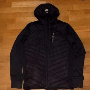  Sail Racing jacka  - Svart hybridjacka från Sail Racing med huva och dragkedja. Jackan har quiltad front och släta ärmar, samt diskret logga på bröstet. Materialmix med polyester och nylon ger en sportig vibe. Perfekt för dig som gillar stilren och funktionell design.