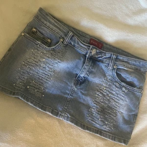 Blå jeanskjol med slitningar från Quiz - Snygg ljusblå jeanskjol från Quiz med slitna detaljer och små glittrande pärlor framtill och på bakfickorna. Stl M-L midja 38cm längd 27 cm •stretchig