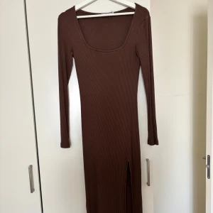 Brun ribbad långklänning från NA-KD - Snygg brun långklänning från NA-KD i ribbad bomullsmix. Klänningen har lång ärm, fyrkantig ringning och en slits framtill som ger en extra edge. Perfekt för dig som gillar en slim och stilren look.