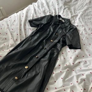 Svart skinnklänning med guldknappar - Snygg svart klänning i skinnimitation från Zara med korta ärmar och stora guldknappar framtill. Klänningen har klassisk krage och rak passform, perfekt för dig som vill sticka ut med en edgy look.