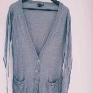 Grå kofta från H&M med knappar strl M - Grå långärmad kofta från H&M i mjuk bomullsblandning. Djup v-ringning, knäppning framtill med stora knappar och två fickor med små rosetter. Perfekt att slänga över en t-shirt eller linne för en chill look.  Helt skick dock nopprig