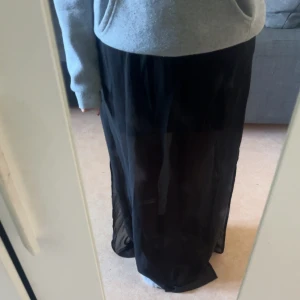 Svart långkjol i transparent tyg - Säljer en svart långkjol i tunt, transparent material som ger en snygg och luftig känsla. Slits på båda sidorna av kjolen. Kjolen har en rak passform och är perfekt för dig som gillar en stilren och enkel look. Passar till både sneakers och boots.