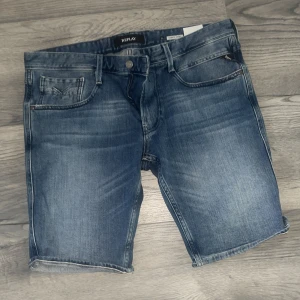Blå jeansshorts från Replay - Snygga blå jeansshorts från Replay med klassisk femficksdesign och slitna detaljer. Shortsen har normal passform, medeltvättad denim och uppvikta benslut. Perfekta för en avslappnad stil under varma dagar.