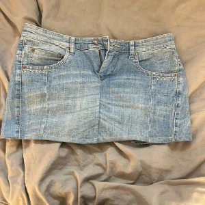 Blå jeanskjol med nitar från MAC - Snygg blå jeanskjol från MAC med kort modell och rå kant nertill. Kjolen har klassiska fickor fram och bak, dekorerade med silvriga nitar för extra edge. Perfekt för dig som gillar en cool och avslappnad stil.