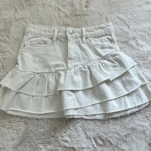 Vit volangkjol i jeans från Gina Tricot - Supersöt vit jeanskjol från Gina Tricot med volanger och råa kanter. Kjolen har klassiska jeansdetaljer som fickor och bälteshällor, och är perfekt för dig som gillar en trendig och lekfull stil. Modellen är kort och har en snygg volangkant som ger extra attityd.
