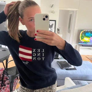 Mörkblå Lexington stickad tröja - Säljer en mörkblå stickad tröja från Lexington med vit text och en rödvit randig flaggdetalj på bröstet. Tröjan har rund halsringning och långa ärmar, perfekt för dig som gillar klassisk preppy stil. Som ny