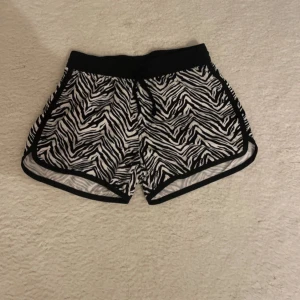 Zebramönstrade sportiga shorts - Snygga shorts med svartvitt zebramönster och elastisk midja. De har svarta kanter och är perfekta för en cool och sportig look. Lätta och luftiga, passar perfekt till sommaren eller träning. Styla med en enkel t-shirt eller hoodie för en avslappnad vibe.
