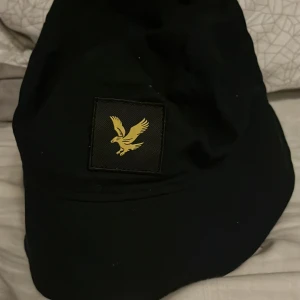 Svart bucket hat från Lyle & Scott - Bucket hat från Lyle & Scott Junior i svart bomull med klassisk gul örnlogga framtill. Hatten har en bred, ljusgrön brätte och är perfekt för en chill och stilren look. Enkel att matcha med streetwear eller casual outfits.