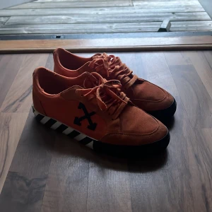 Orange Off-White sneakers strl 41 - Snygga orange sneakers från Off-White med svart logga på sidan och coola svartvita ränder på sulan. Skorna har snörning och är gjorda i canvas och mocka. Unik detalj med röd plasttag på sidan och tryckt text. Perfekta för dig som gillar streetwear och vill sticka ut. Nypris runt 3000kr. PRIS KAN DISKUTERAS.