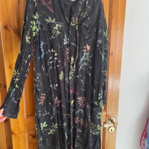Svart blommig långklänning Dedicated - Långklänning från Dedicated i svart med färgglatt blommönster. Klänningen har långa ärmar, knappar framtill och två bröstfickor. Tillverkad i mjukt Tencel™ lyocell-material som känns svalt och skönt.