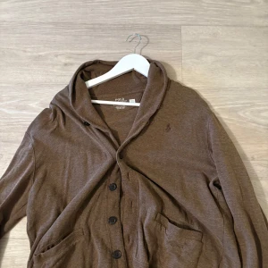 Brun kofta med huva från Polo Ralph Lauren - Snygg brun kofta från Polo Ralph Lauren med huva och knäppning framtill. Tröjan har två fickor, långa ärmar och är tillverkad i mjuk bomull. Perfekt för dig som gillar en chill och avslappnad stil.